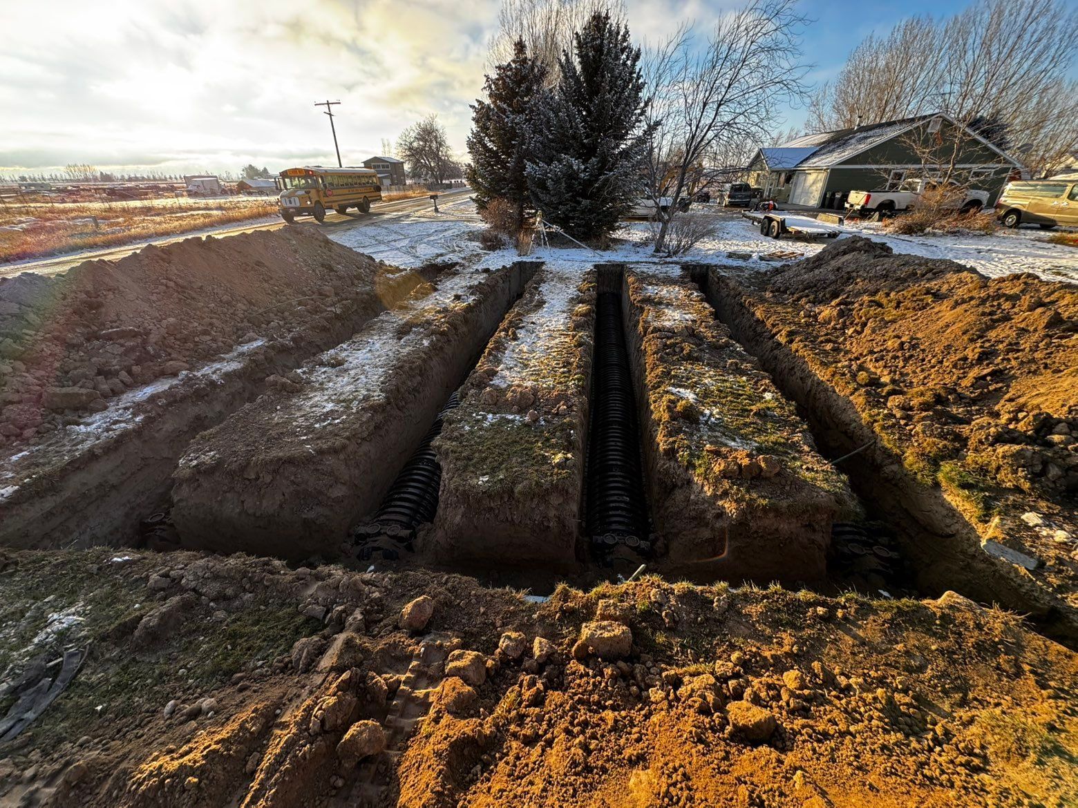 Three deep, parallel trenches cut into a snow-dusted, grassy residential yard with a school bus visible in the distance. 