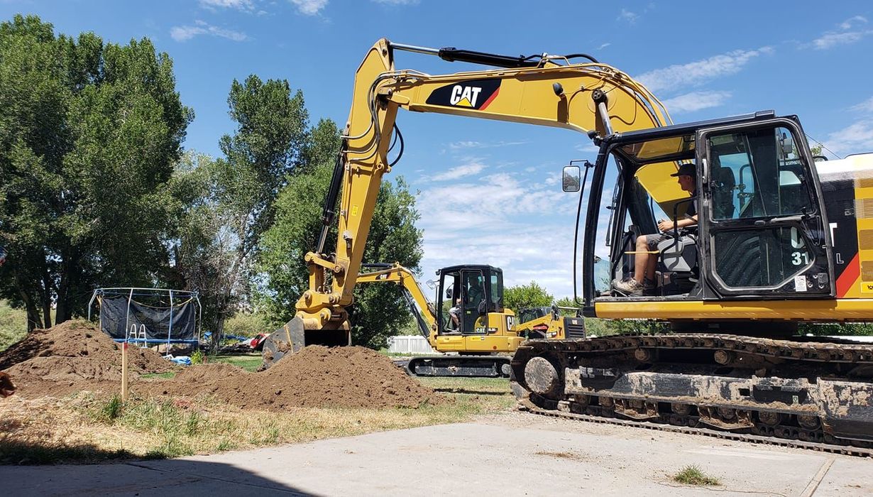Two yellow CAT excavators operate in a sunny, tree-lined yard with a pile of excavated dirt.