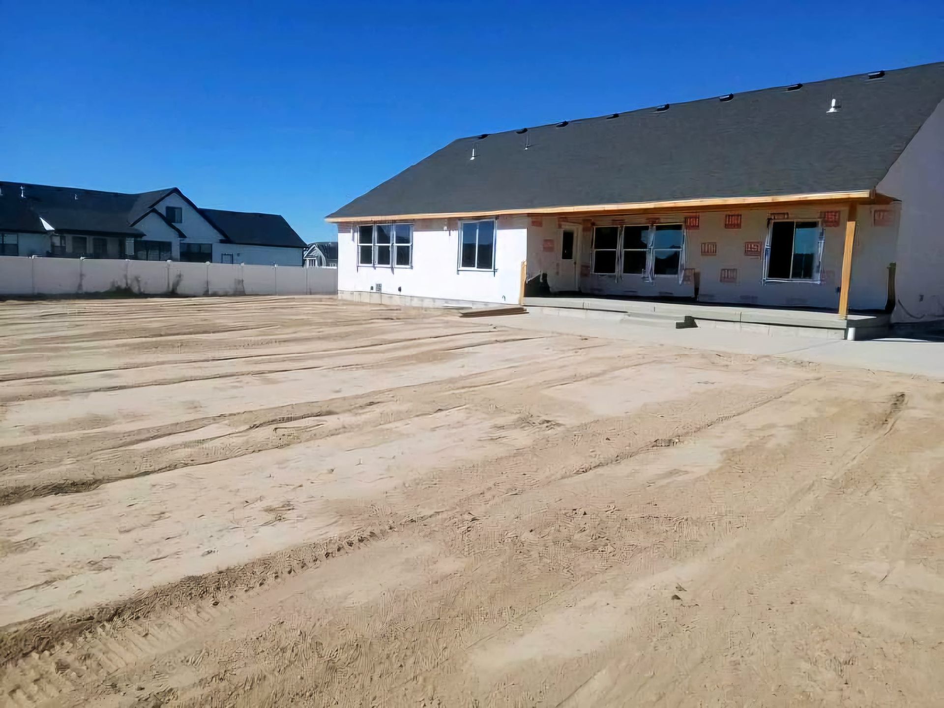 A newly built house with a dark roof and white exterior, featuring a large, dirt-covered backyard under a clear blue sky. 