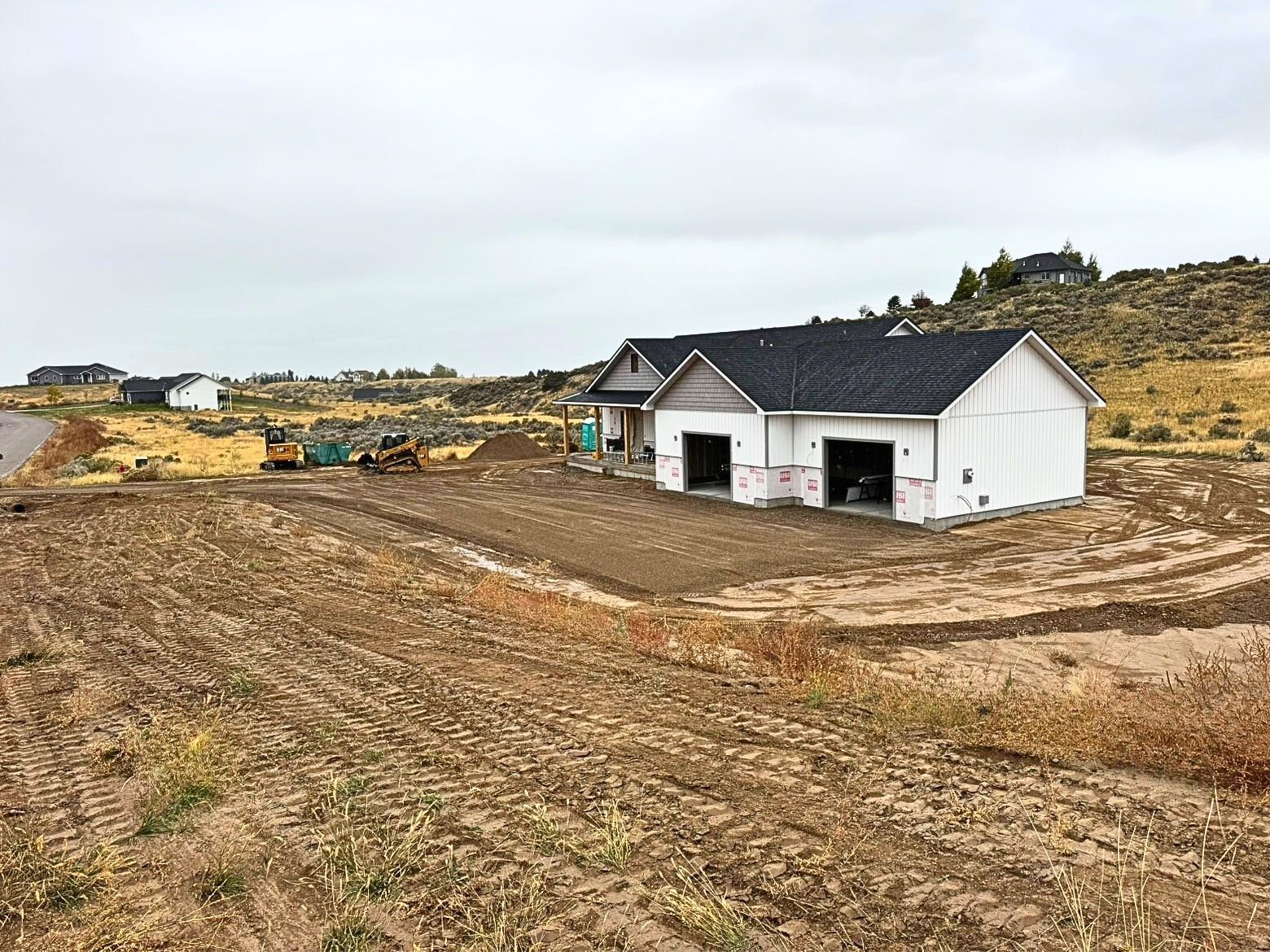 A white, newly constructed house with a dark shingled roof stands on a graded dirt lot under an overcast sky. 