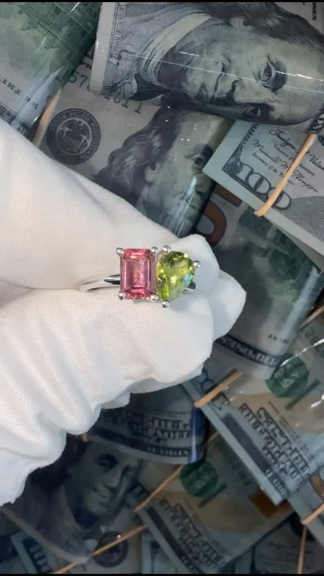A gloved hand holds a pink and green gemstone ring over scattered U.S. dollar bills.
