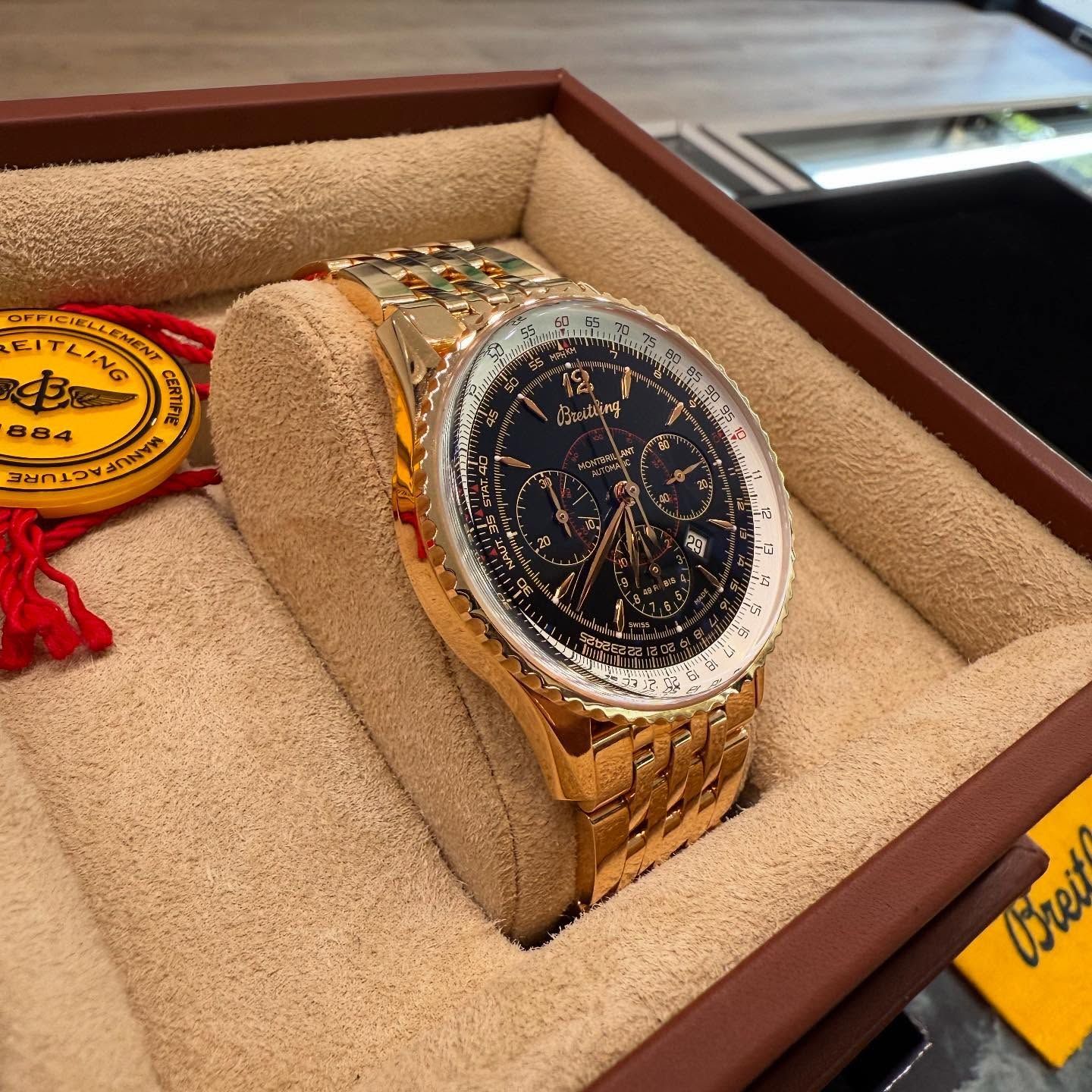 Gold wristwatch with black dial in a tan display box, shown at an angle on a wooden surface.
