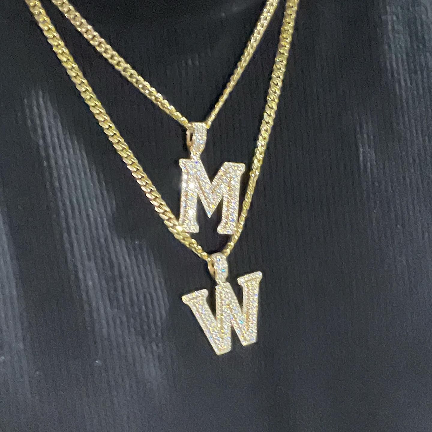 Two gold chains with diamond pendants shaped like M and W on a black shirt