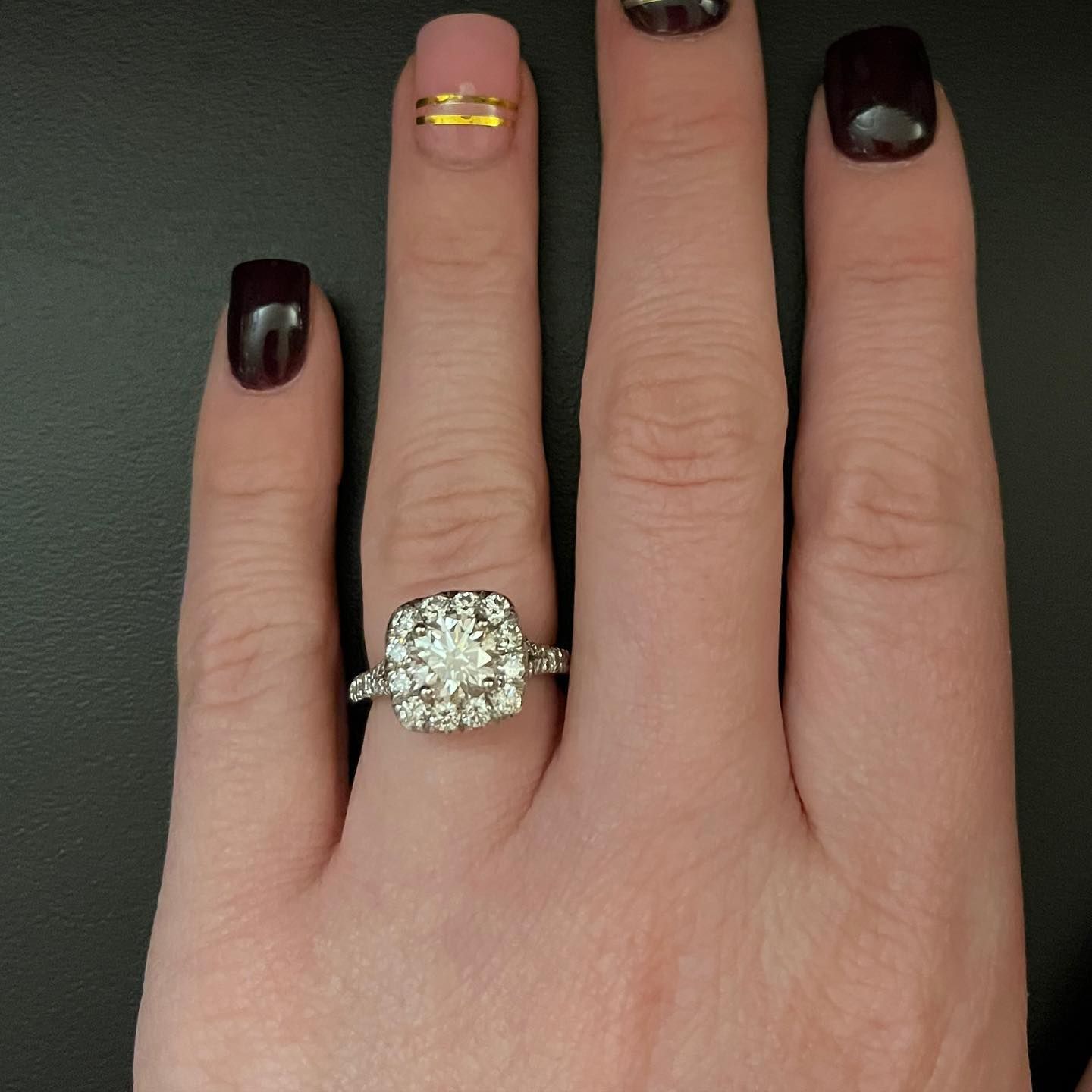 Hand with dark nail polish wearing a large diamond ring and gold band on a gray background