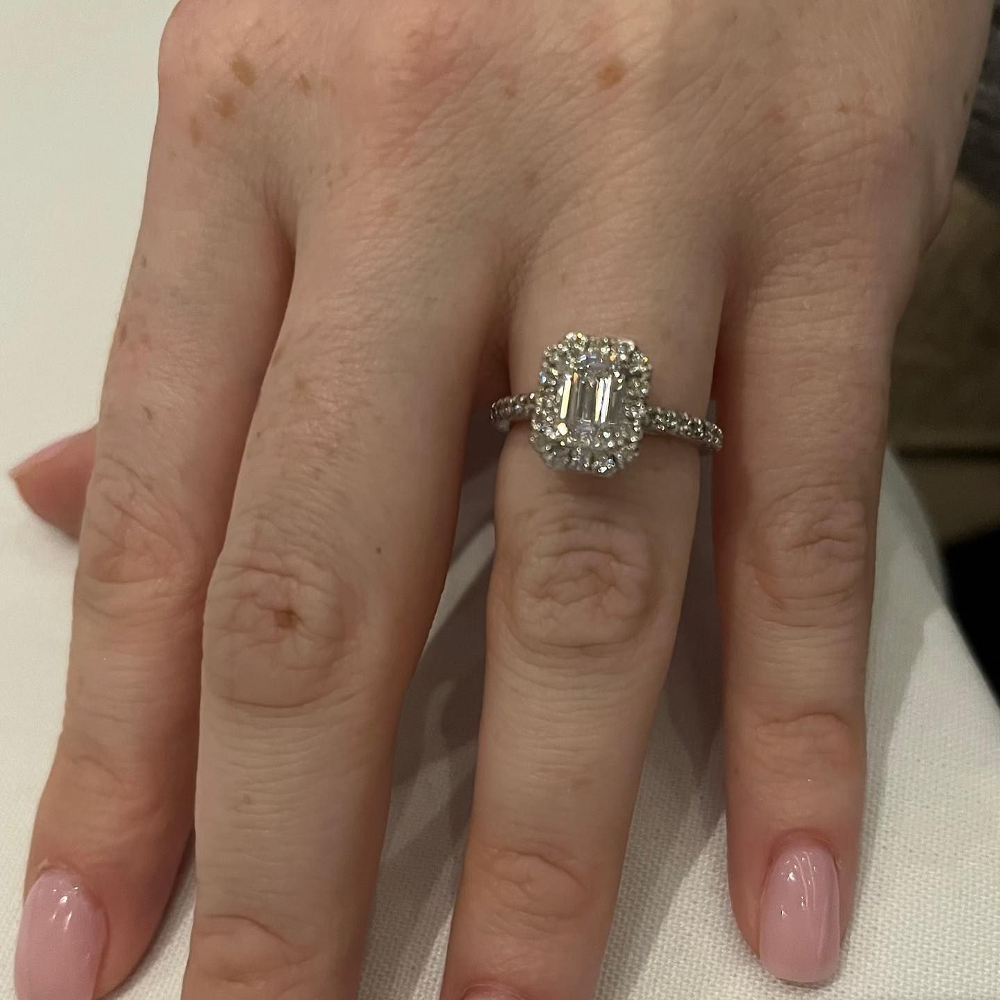 Hand with pink manicured nails wearing a large rectangular diamond ring on a white surface