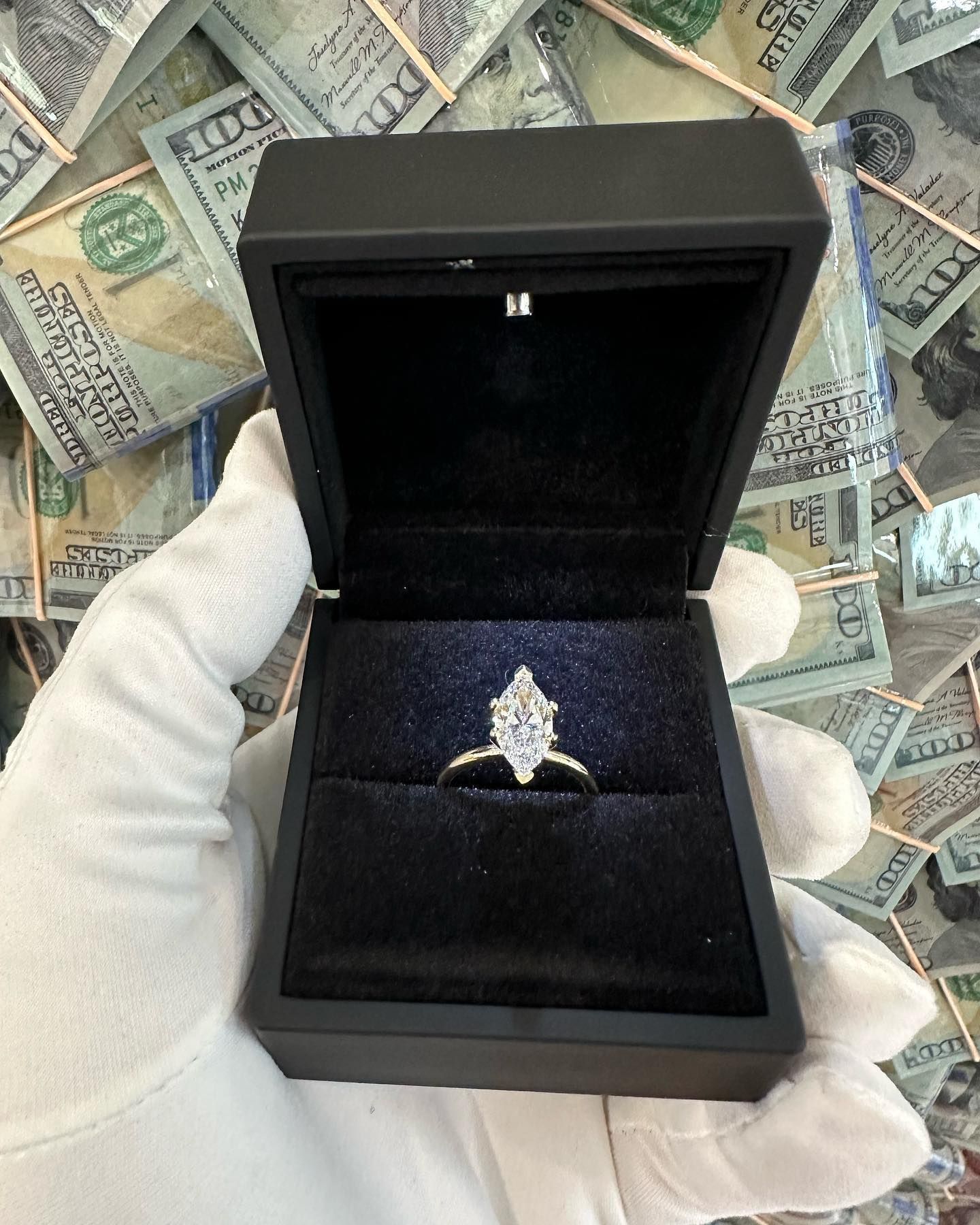 White-gloved hand holding open black ring box with a diamond ring inside, on a money-print background