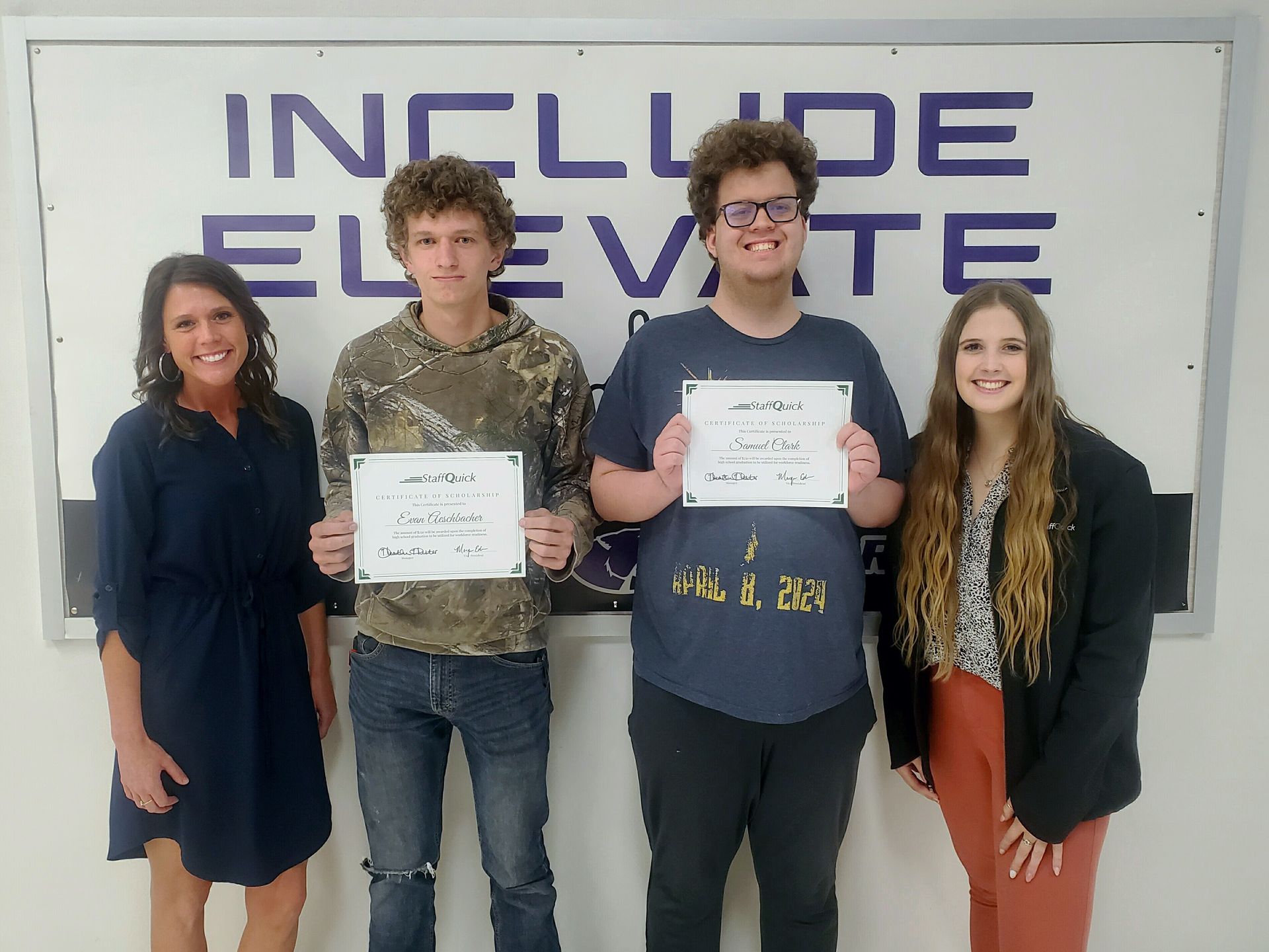 Four people stand in front of a sign. Two young men hold certificates. Two women stand on either side.