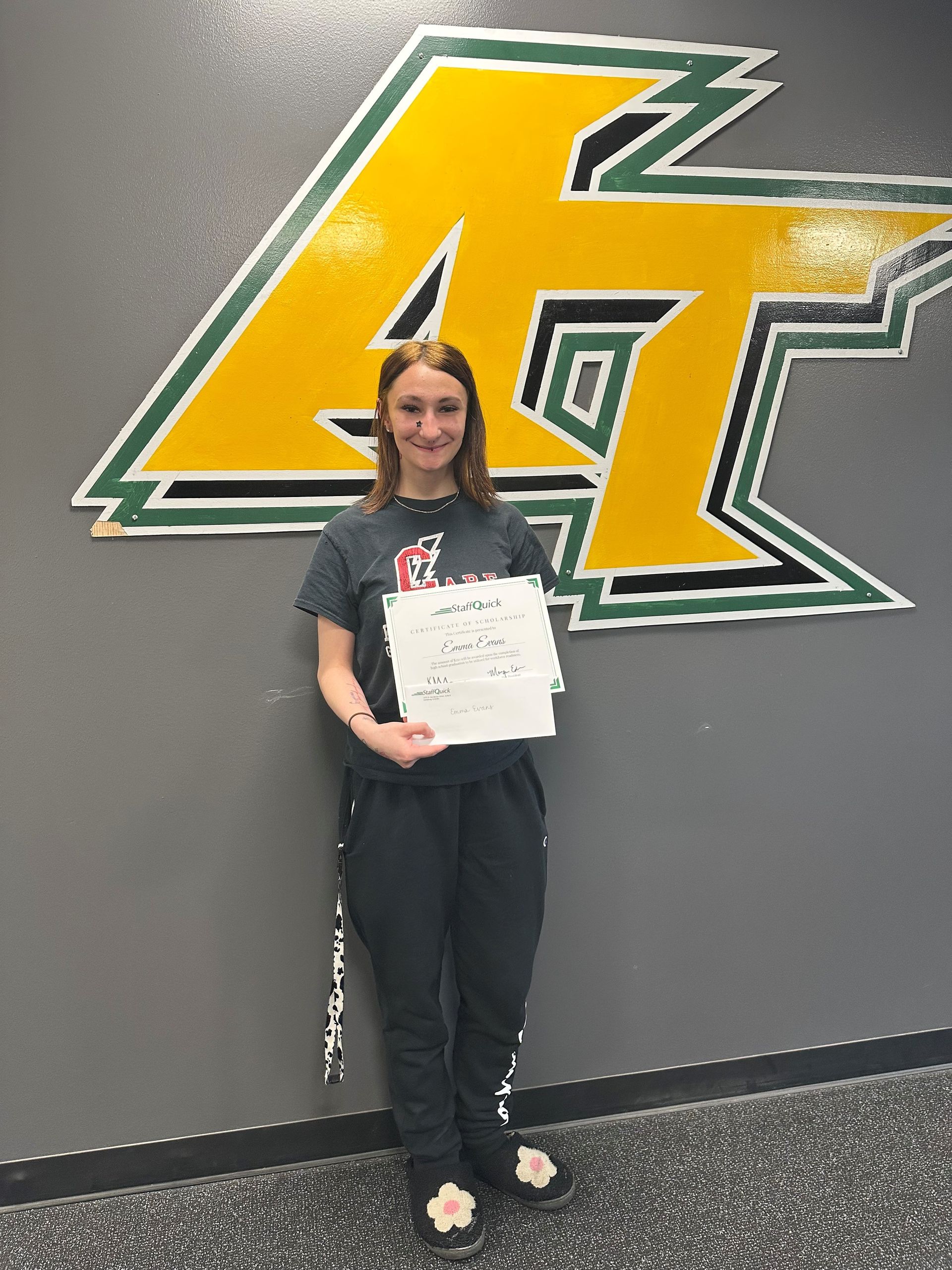 Woman standing in front of a logo holding paper, wearing black pants and shirt.