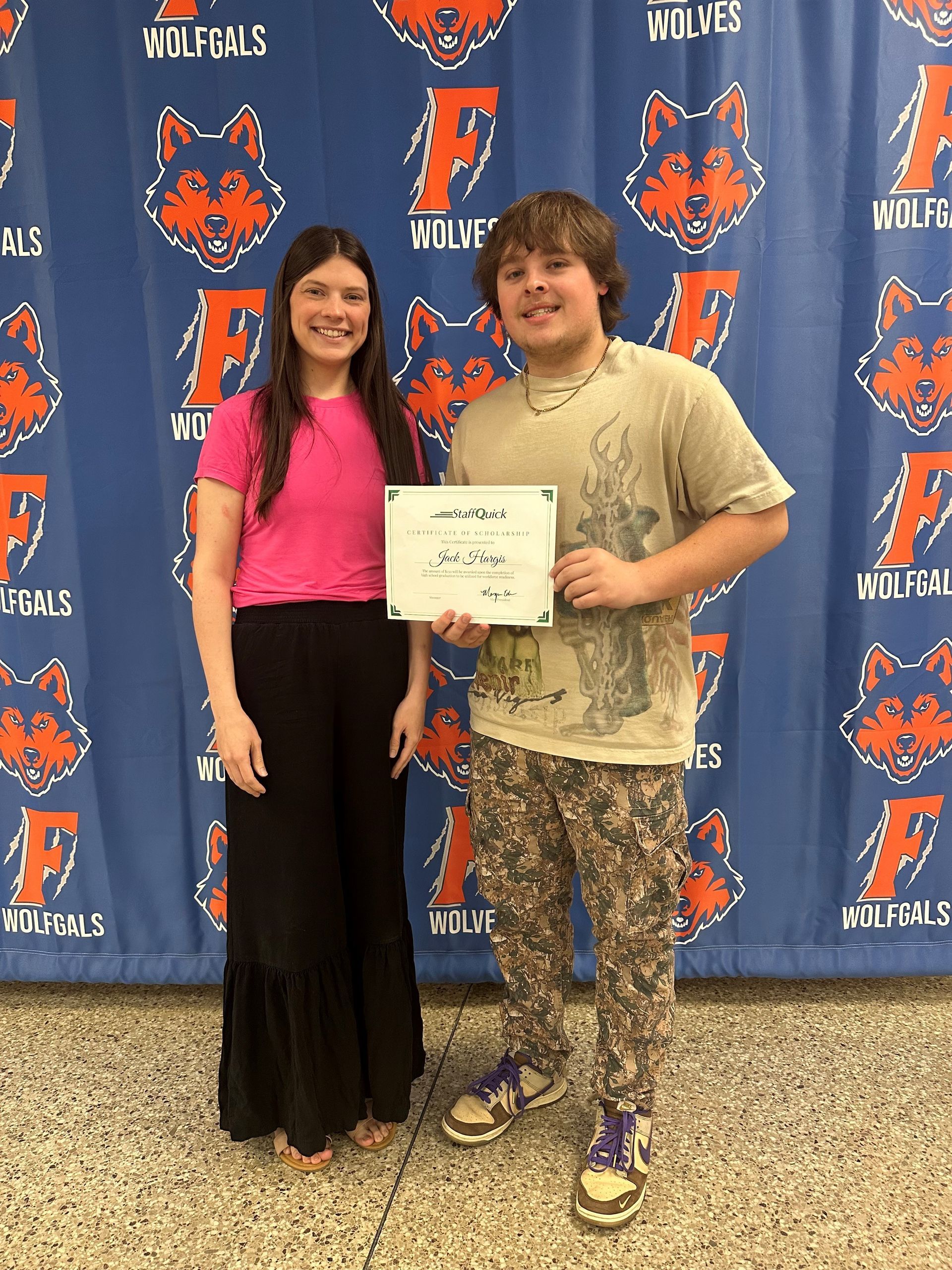 Woman and boy holding a certificate, smiling, in front of a school banner.