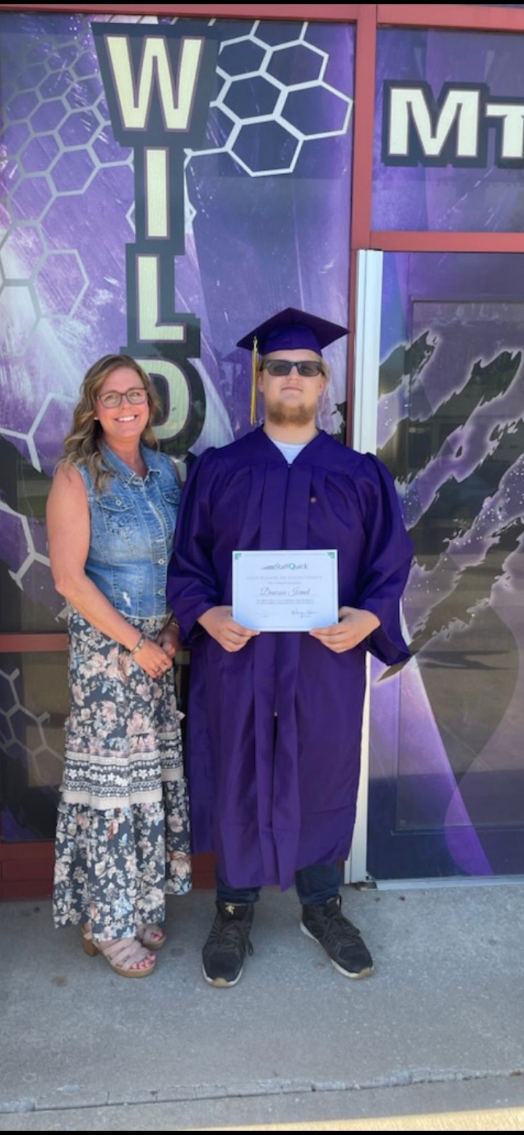 A graduate in a purple gown and cap holding a diploma, standing with a woman in front of a