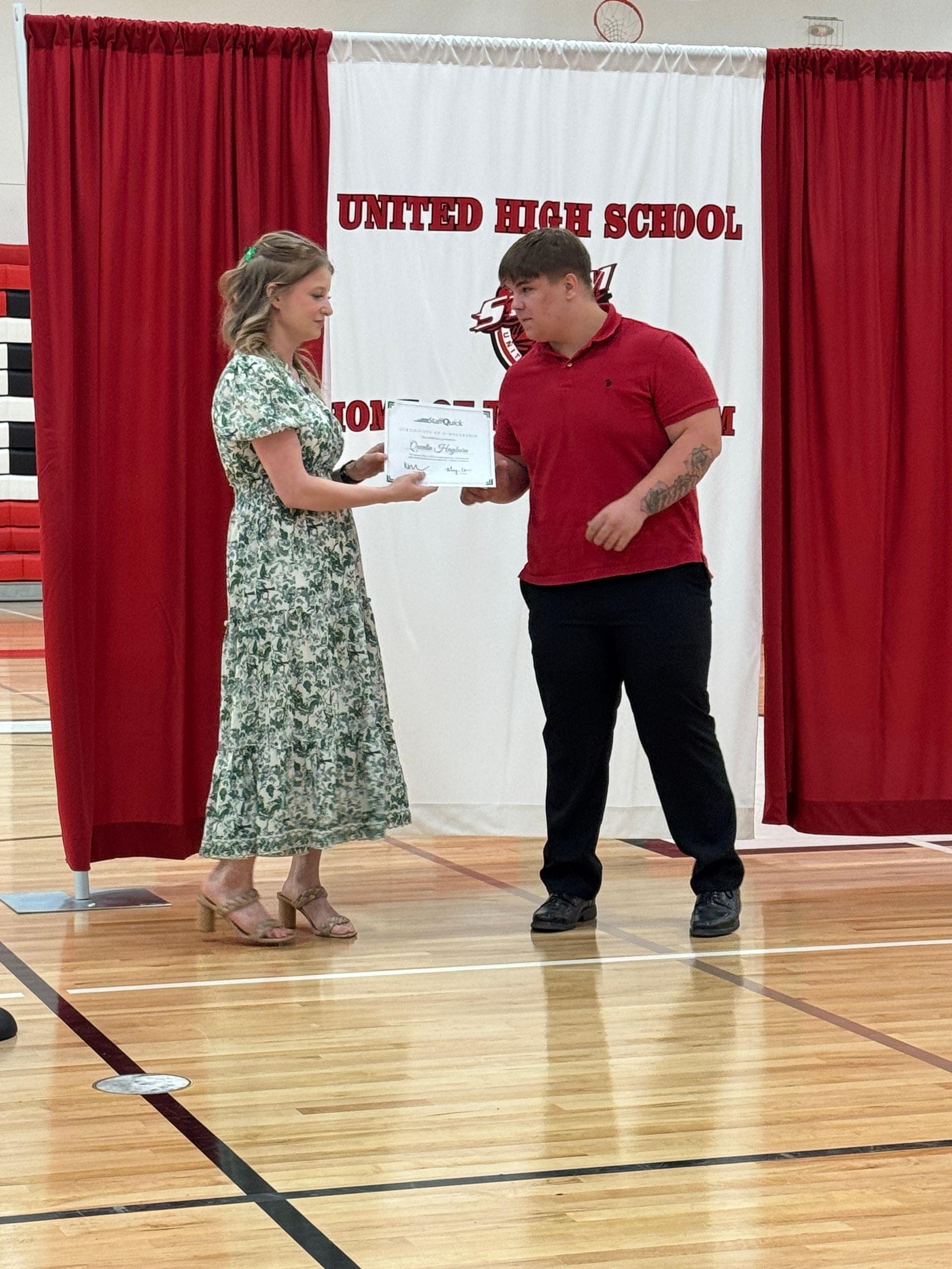 Woman presents a certificate to a man at United High School, red curtains in background.