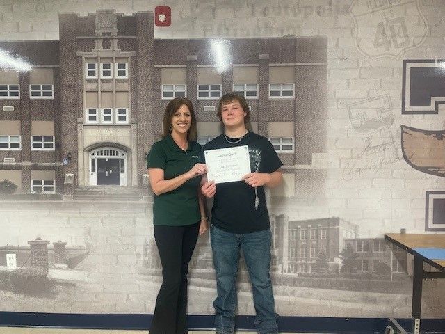 Woman in green shirt presents certificate to a teen in front of school mural.