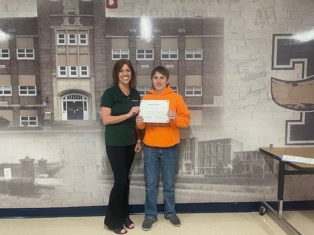 Woman in green shirt and boy in orange hoodie holding certificate in front of school mural.