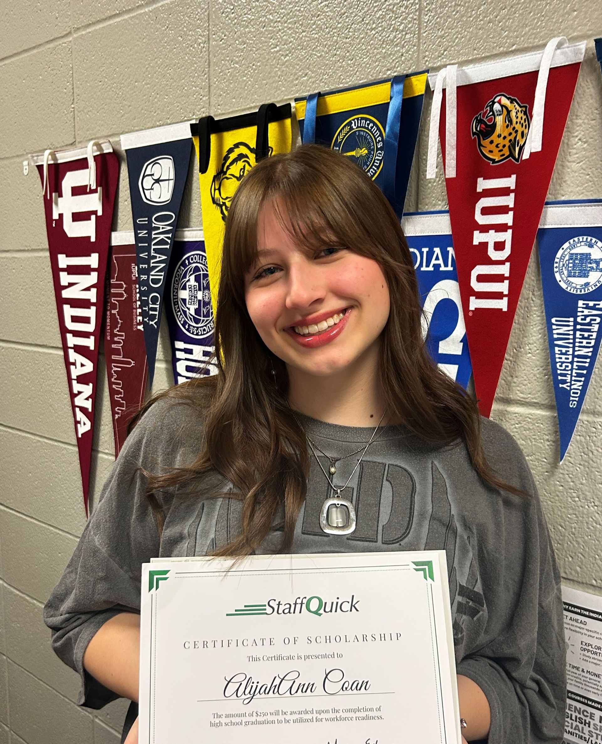 Smiling young woman holds certificate in front of college pennants, brick wall background.