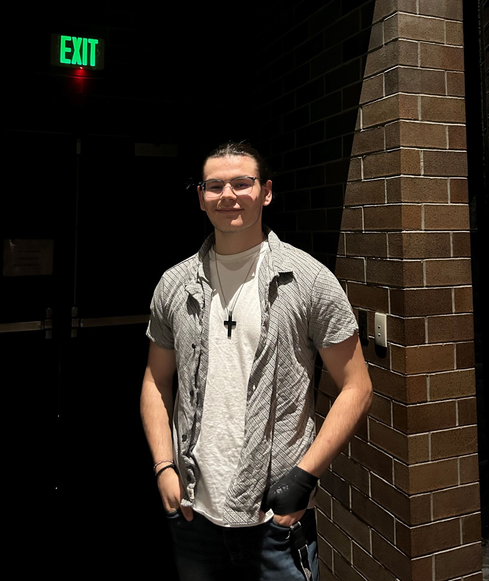 Young man in casual clothes standing near a brick wall and an exit sign, smiling slightly.