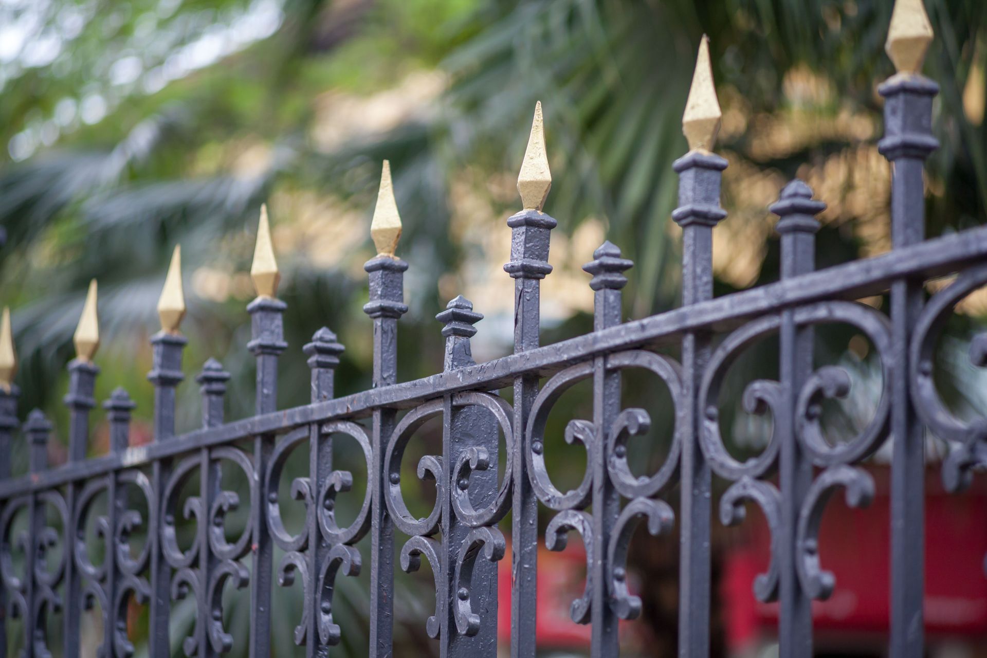 Black wrought iron fence with gold-tipped points, ornate swirls, and blurred green foliage in the background.
