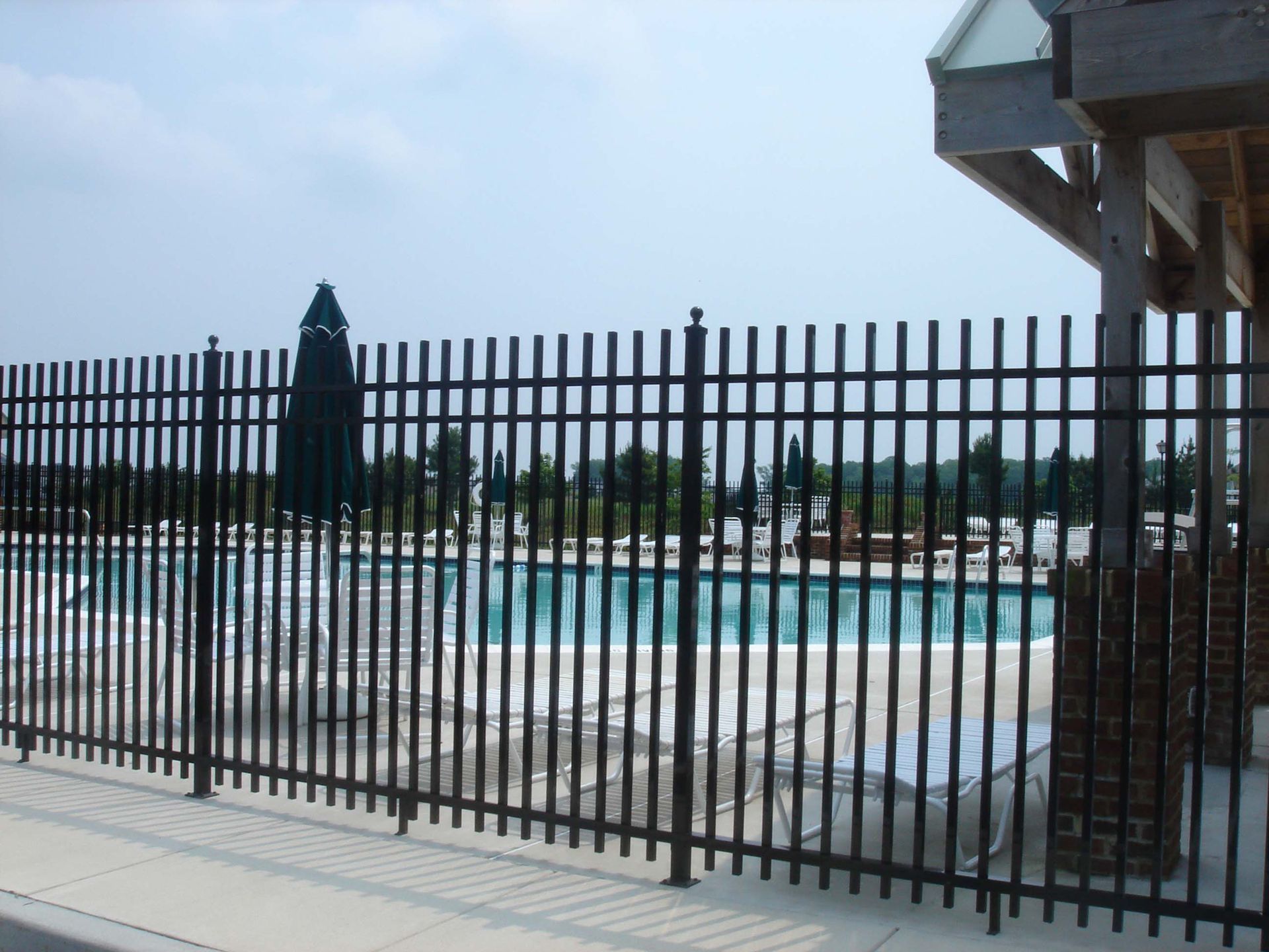 Black metal fence bordering a pool area. An umbrella and trees are in the background. Blue sky overhead.