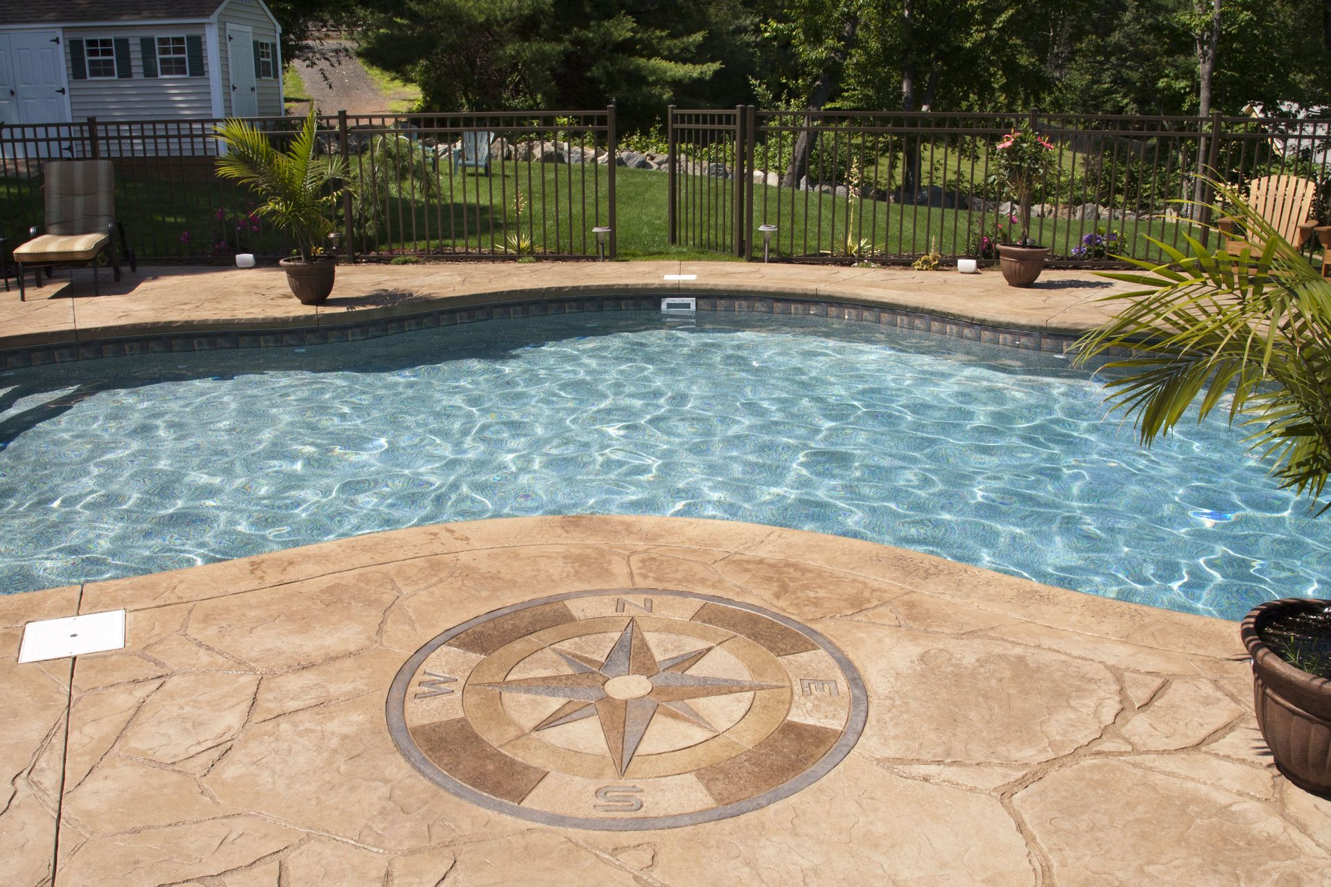 Swimming pool with decorative compass design on the stone patio. Surrounding fence, trees, and house are in the background.