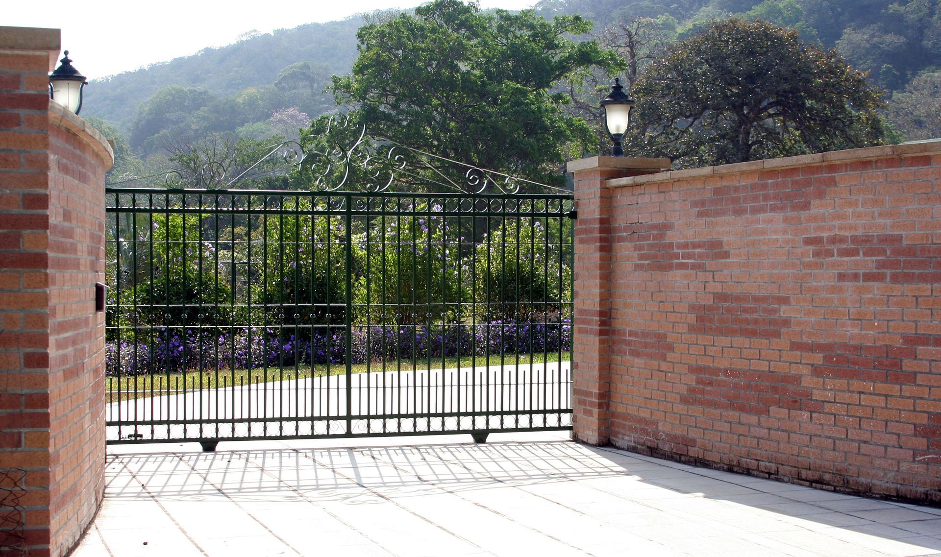 Iron gate between brick pillars with lanterns, opening to a garden with a mountain in the background.