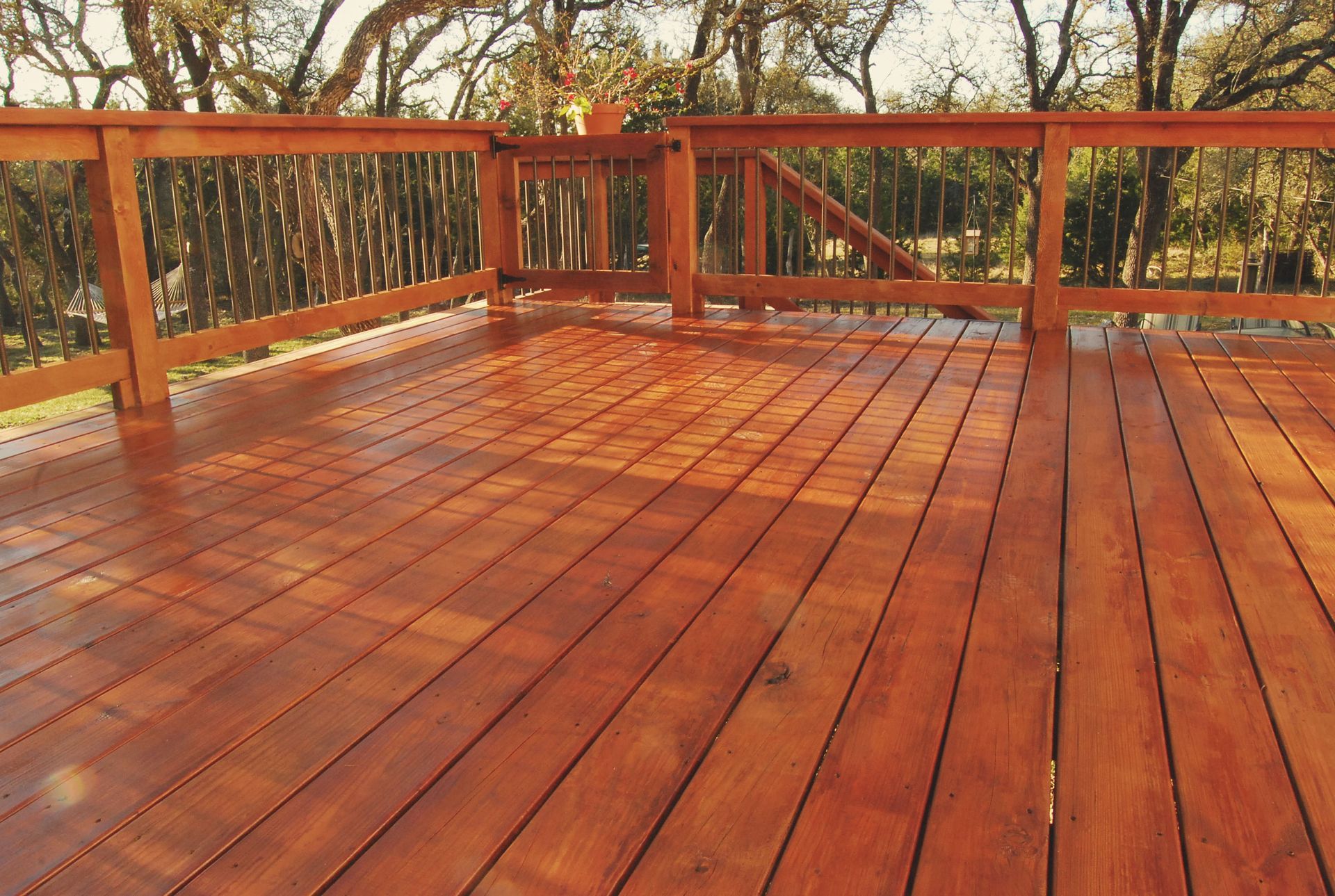 Wooden deck with railing. Shiny, reddish-brown boards, surrounded by a wooden railing and trees in the background.
