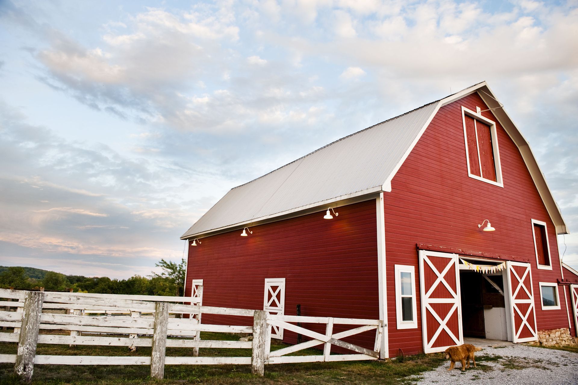 Red barn with white doors and roof, set against a cloudy sky, with a white fence in front.
