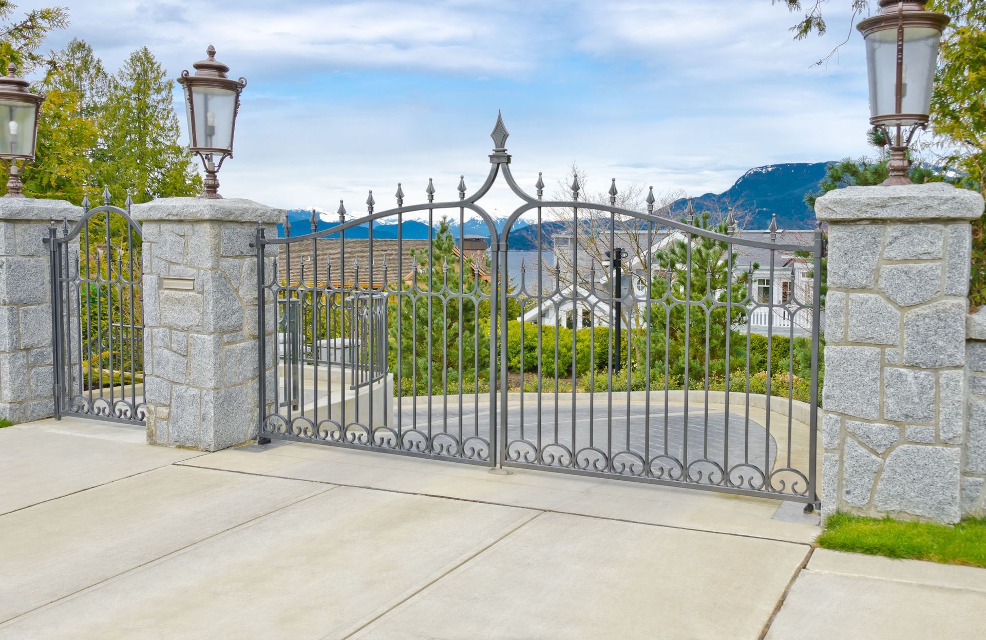 Stone pillars with iron gates and lamps along a concrete driveway, a house visible in the background.