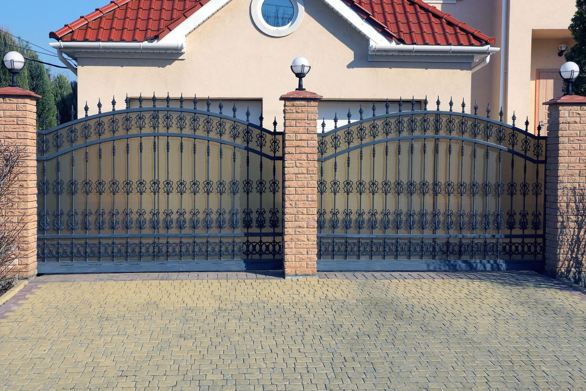 Ornamental iron gates at the entrance of a house, brick pillars, cobblestone driveway.