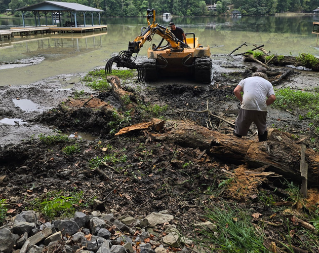 Yellow excavator clearing muddy riverbank while a man stands nearby beside the water