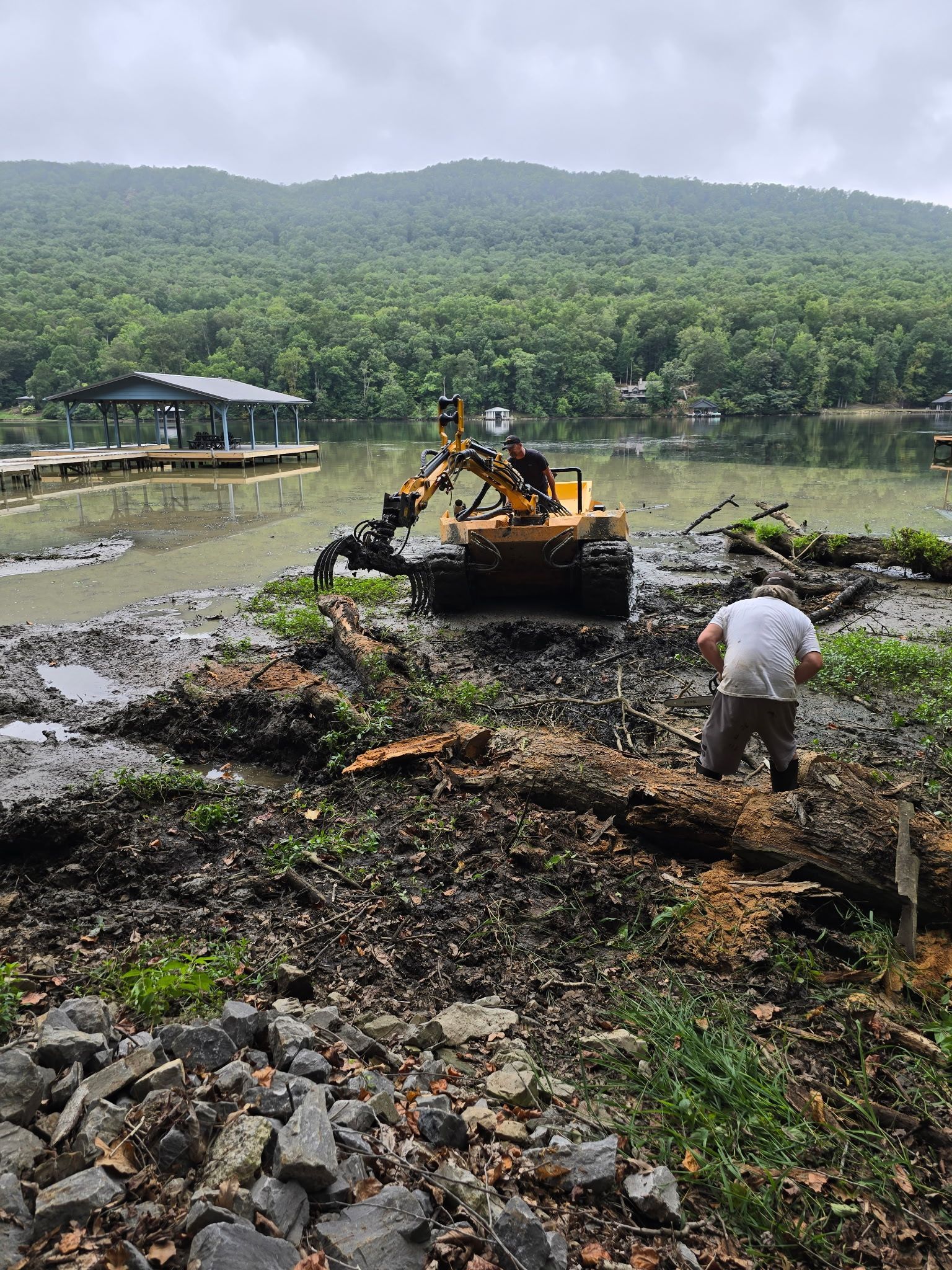 Bulldozer clearing debris by a lakeside, with a worker nearby and forested hills in the background