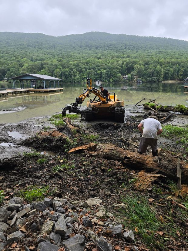 Bulldozer clearing muddy lakeshore debris with a worker nearby, forested hills and water in the background