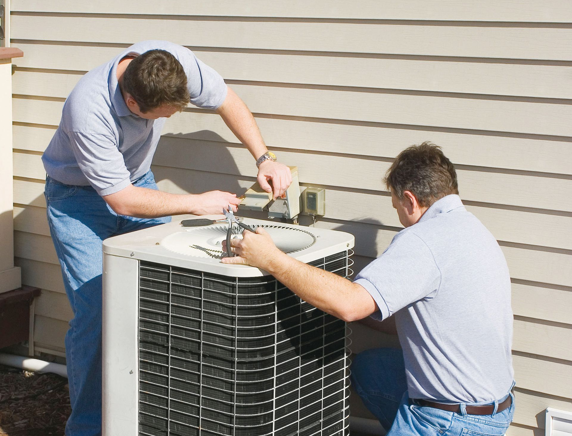 Two men repairing an outdoor air conditioning unit next to a light-colored building.