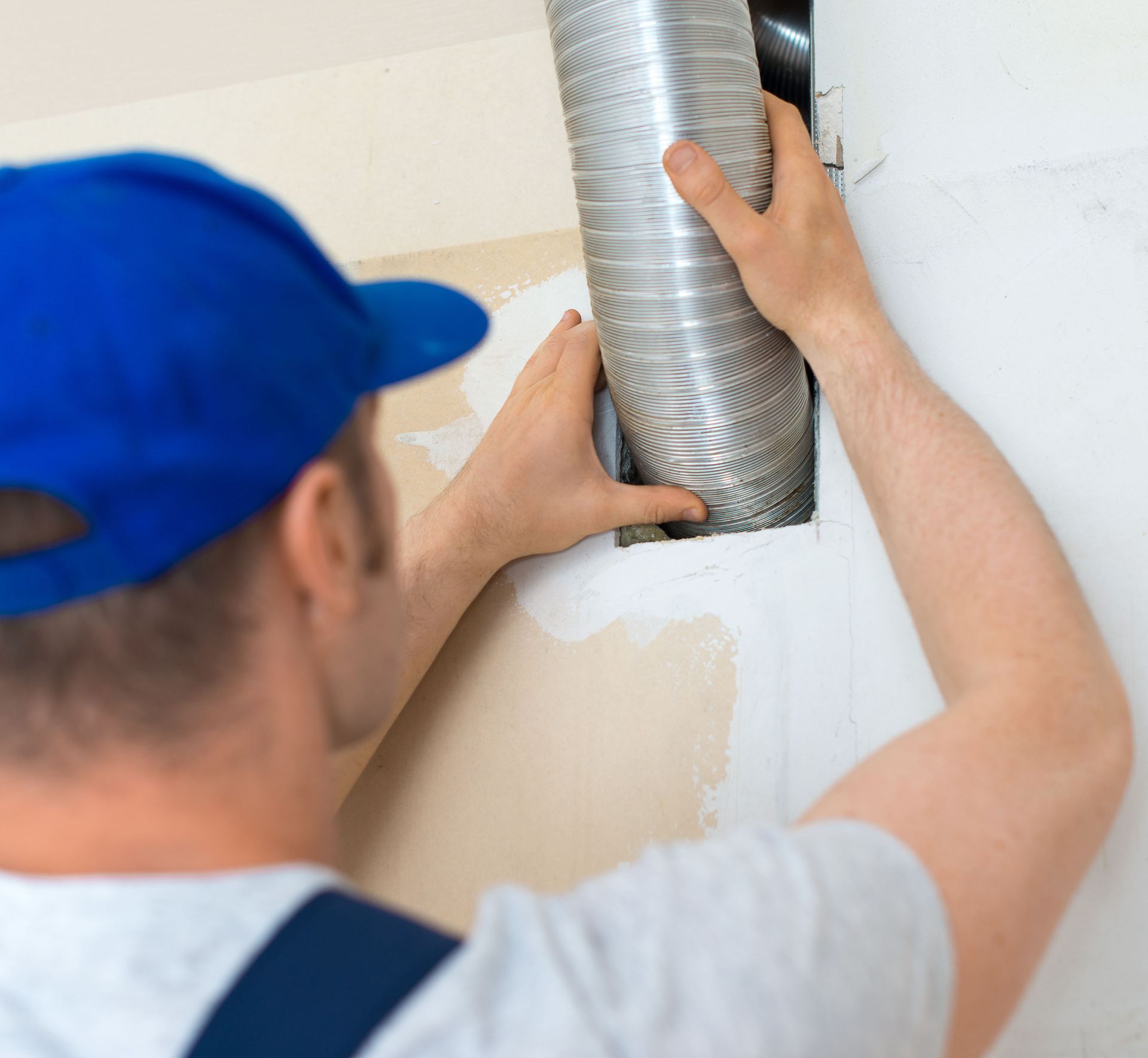 Person in blue cap connecting flexible metal ductwork to an opening in a white wall.