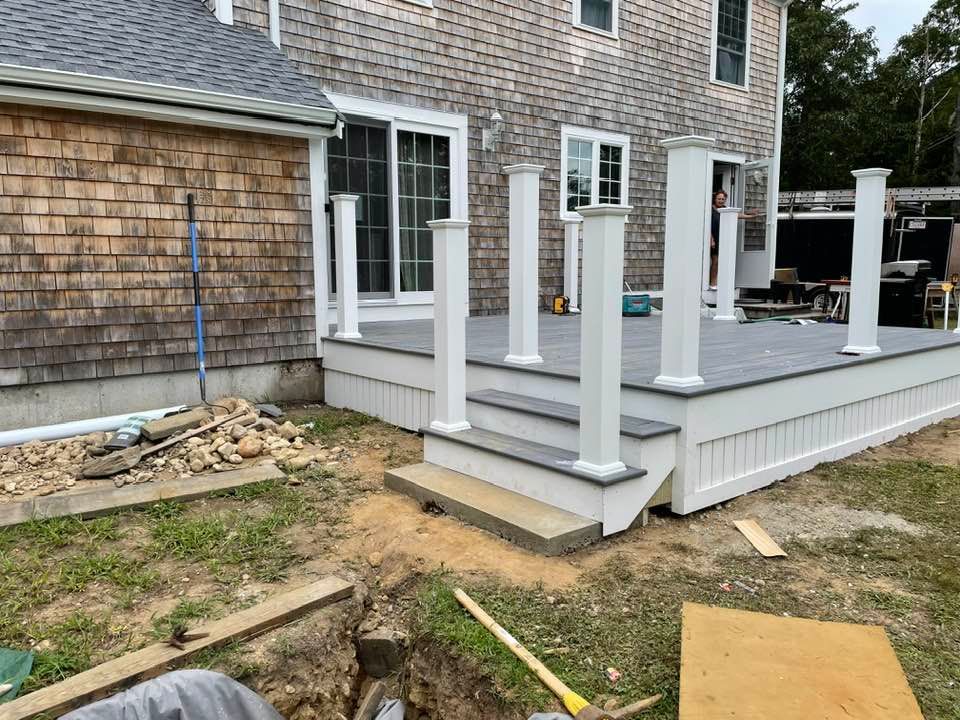 Deck with white pillars and steps, next to a house with brick siding and a sliding glass door.