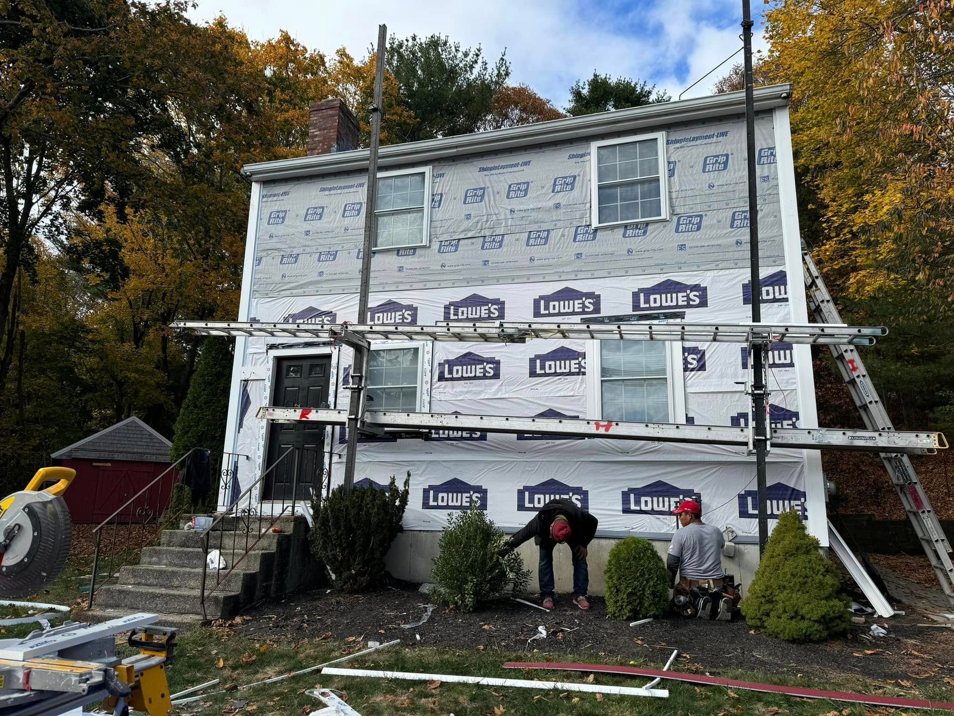 Two story house under renovation, workers on scaffolding, covered in blue wrap, overcast day.