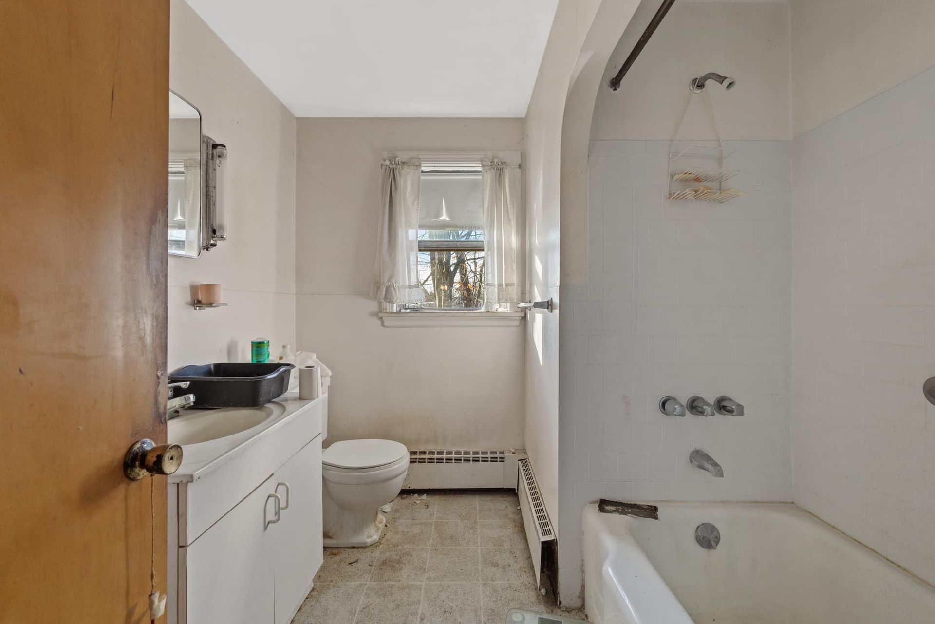 A small bathroom with a toilet, sink, and bathtub. Off-white walls, window, and a wooden door.