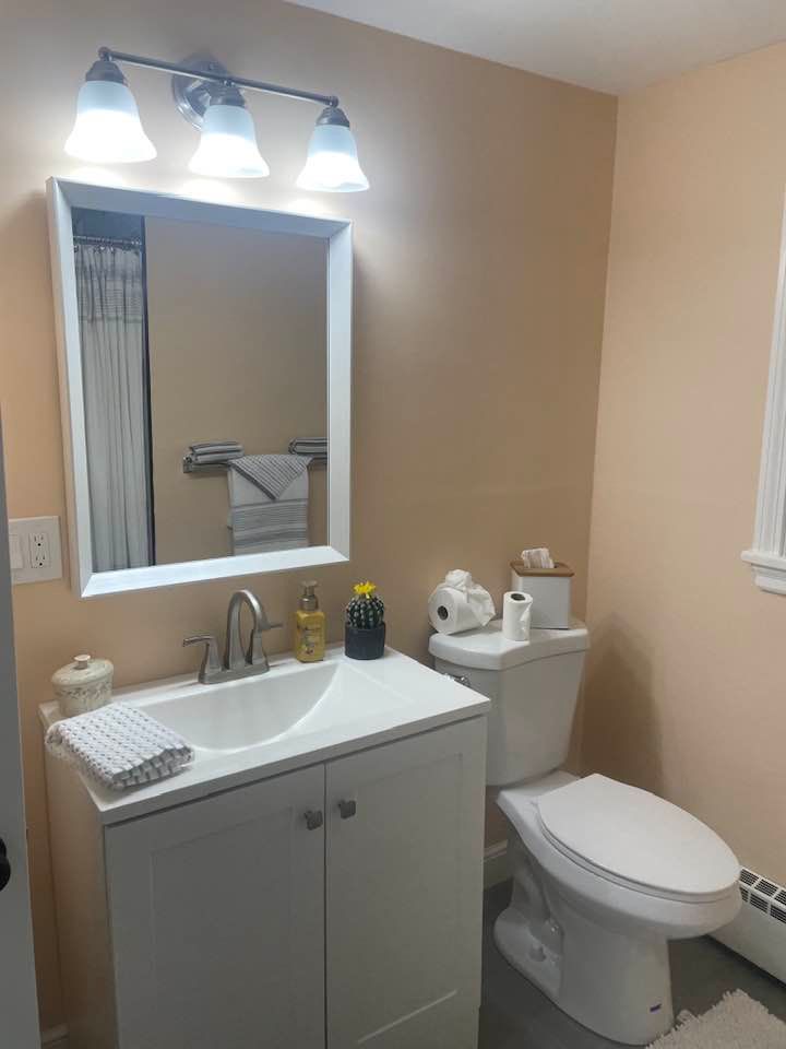 A small bathroom with white vanity, toilet, and peach-colored walls. A mirror and light fixture are above the sink.
