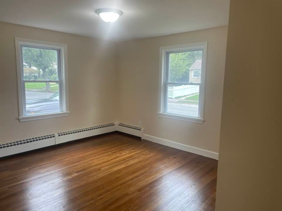 Empty bedroom with hardwood floors, two windows, and beige walls.