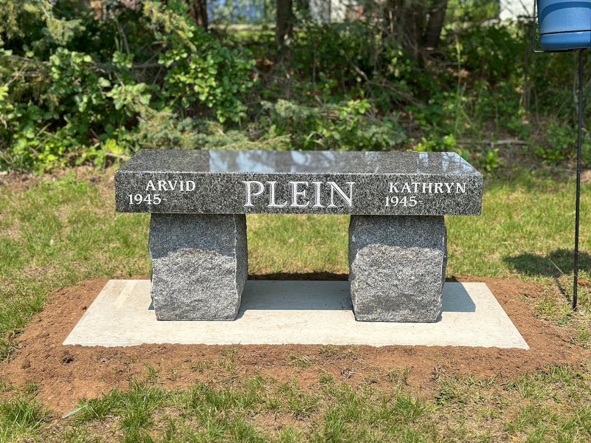A granite bench memorial in a grassy area, engraved with 