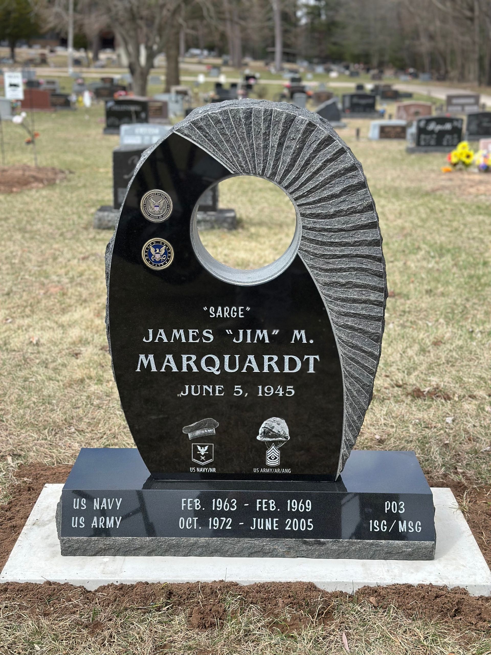 A black granite headstone in a cemetery with a central circular cutout, engraved with the name James M. Marquardt.