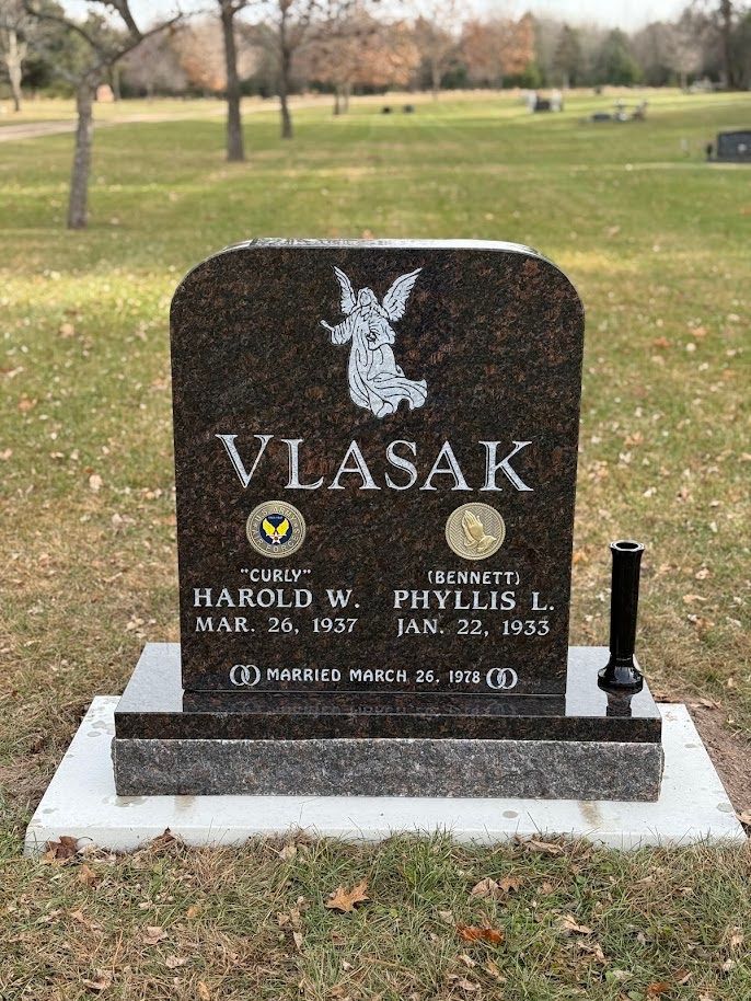 A Vlasak family gravestone featuring an angel carving, names, dates, and a metal flower vase in a cemetery.