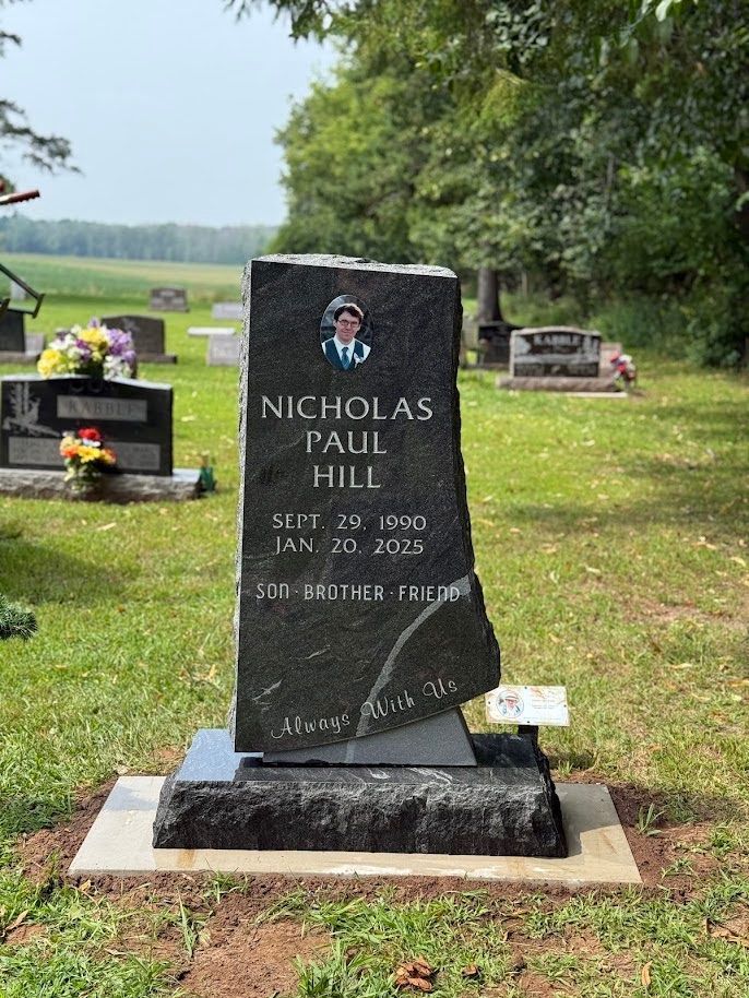 A black, upright granite gravestone for Nicholas Paul Hill, standing in a grassy cemetery.