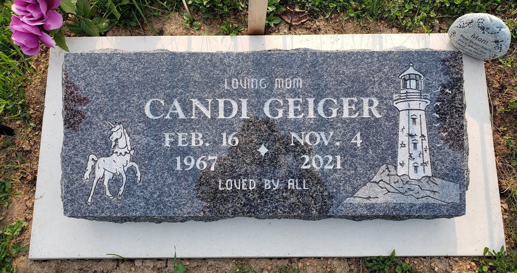 A dark gray rectangular granite headstone for Candi Geiger (1967–2021) with carvings of a horse and a lighthouse.