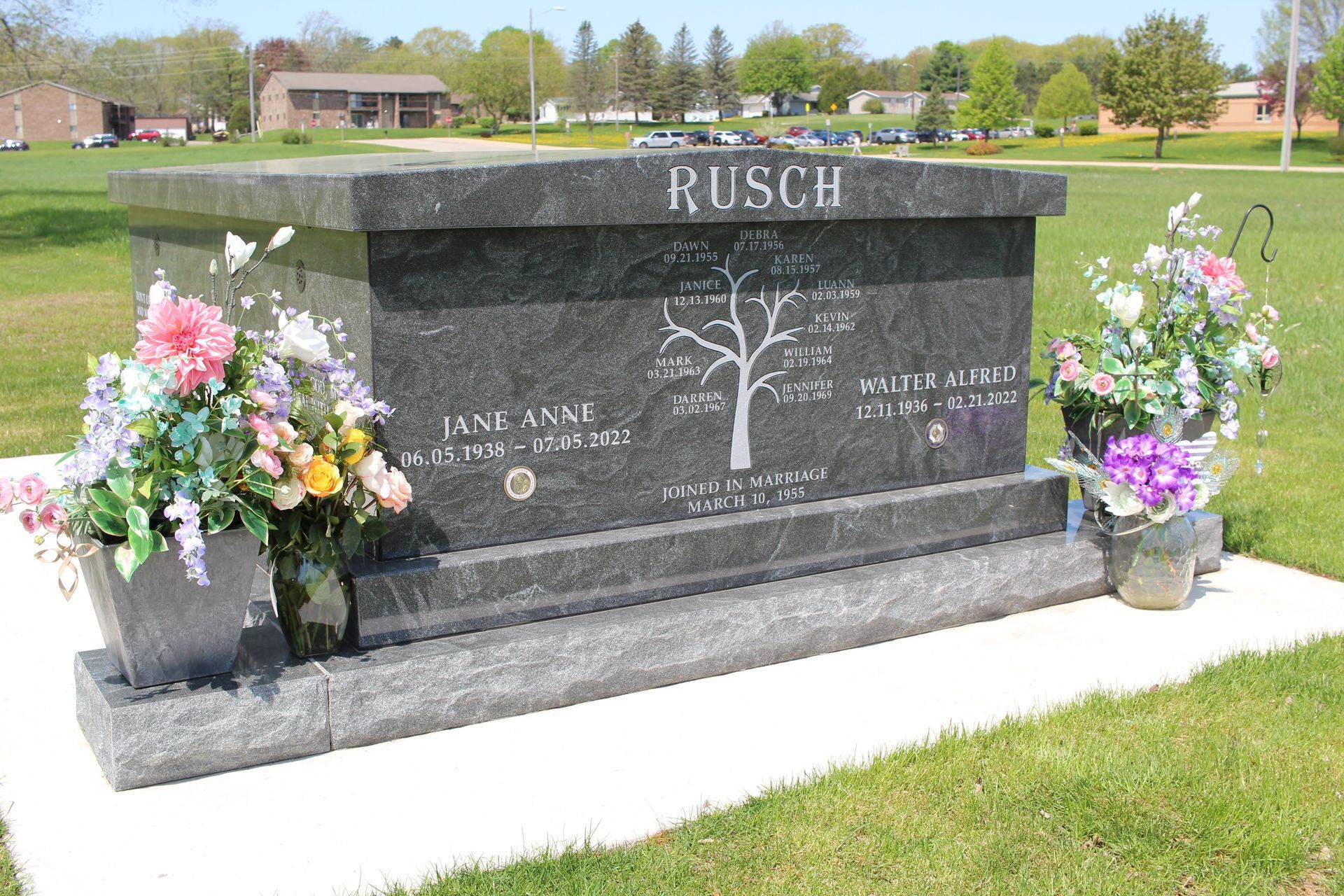 A dark granite Rusch family gravestone with a tree carving and two flower arrangements on a grassy lawn.