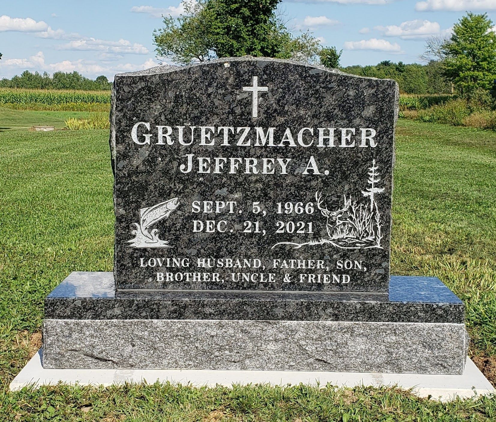 A black granite headstone for Jeffrey A. Gruetzmacher (1966–2021) with engraved fish and deer imagery, set in a field.