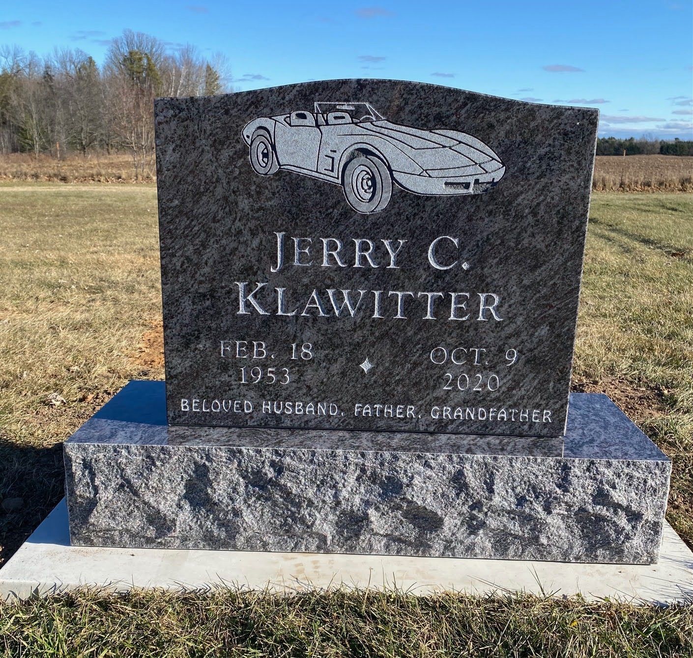 A black granite headstone with an engraved sports car, name Jerry C. Klawitter, dates, and family roles in a grassy field.