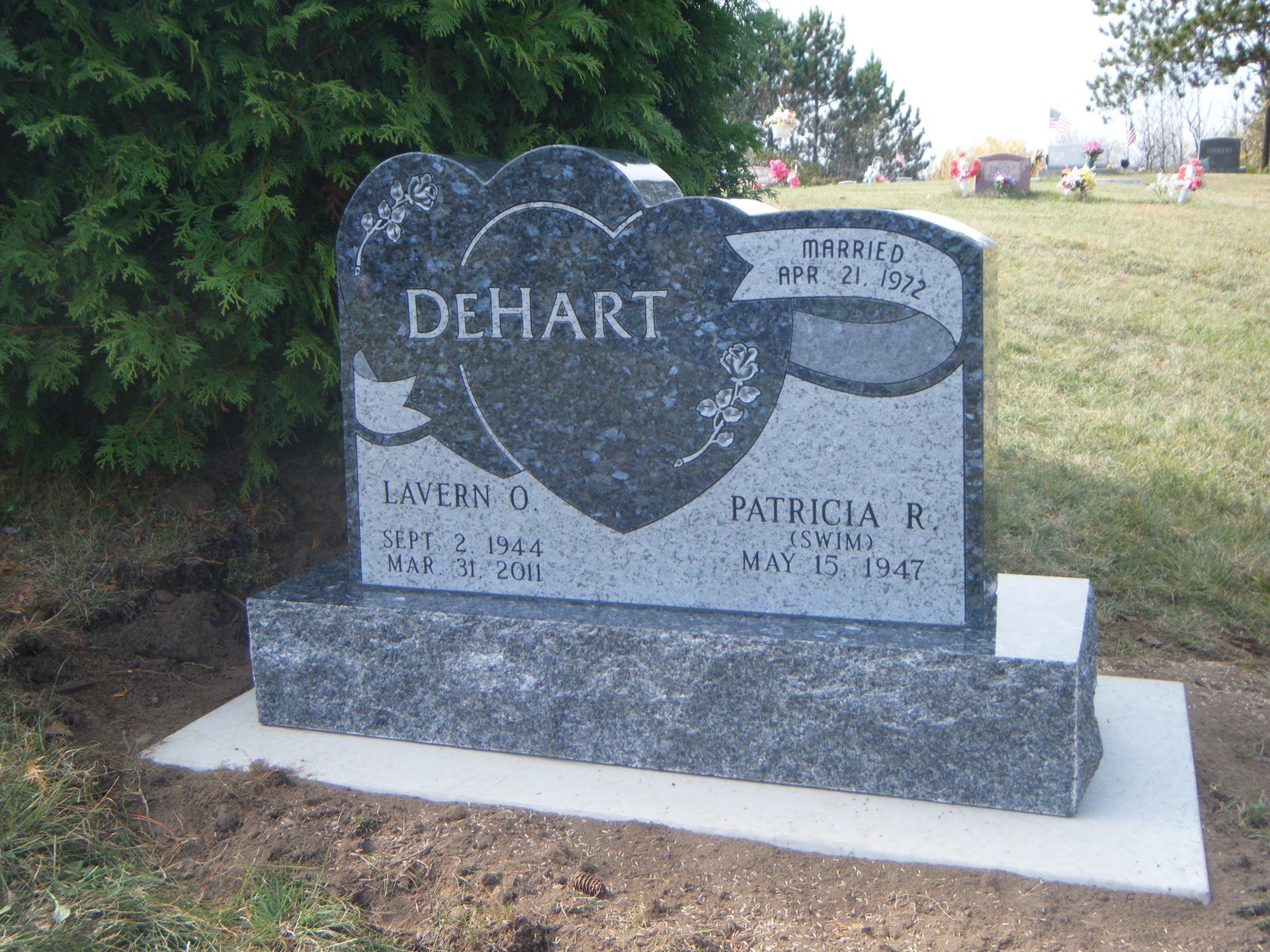 A gray granite DeHart family tombstone with heart engravings, situated on a concrete base in a grassy cemetery.