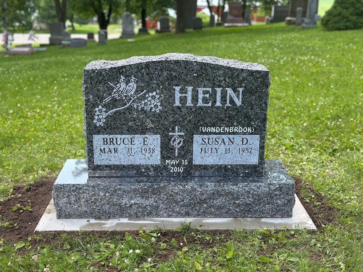 A dark grey granite upright headstone for the Hein family, featuring names and dates, centered in a grassy cemetery.