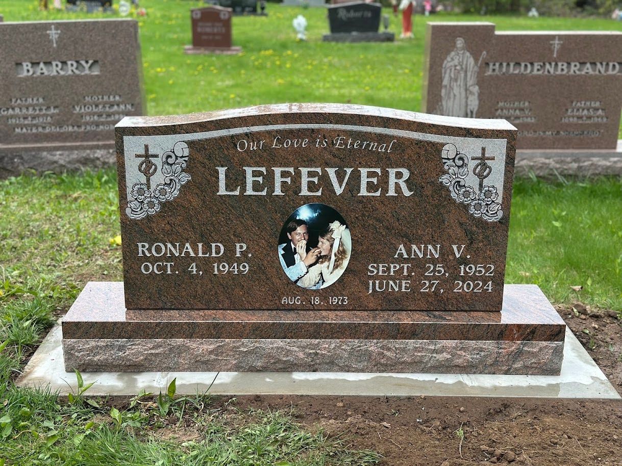 A brown granite headstone for Ronald P. and Ann V. Lefever, centered in a cemetery with surrounding markers.