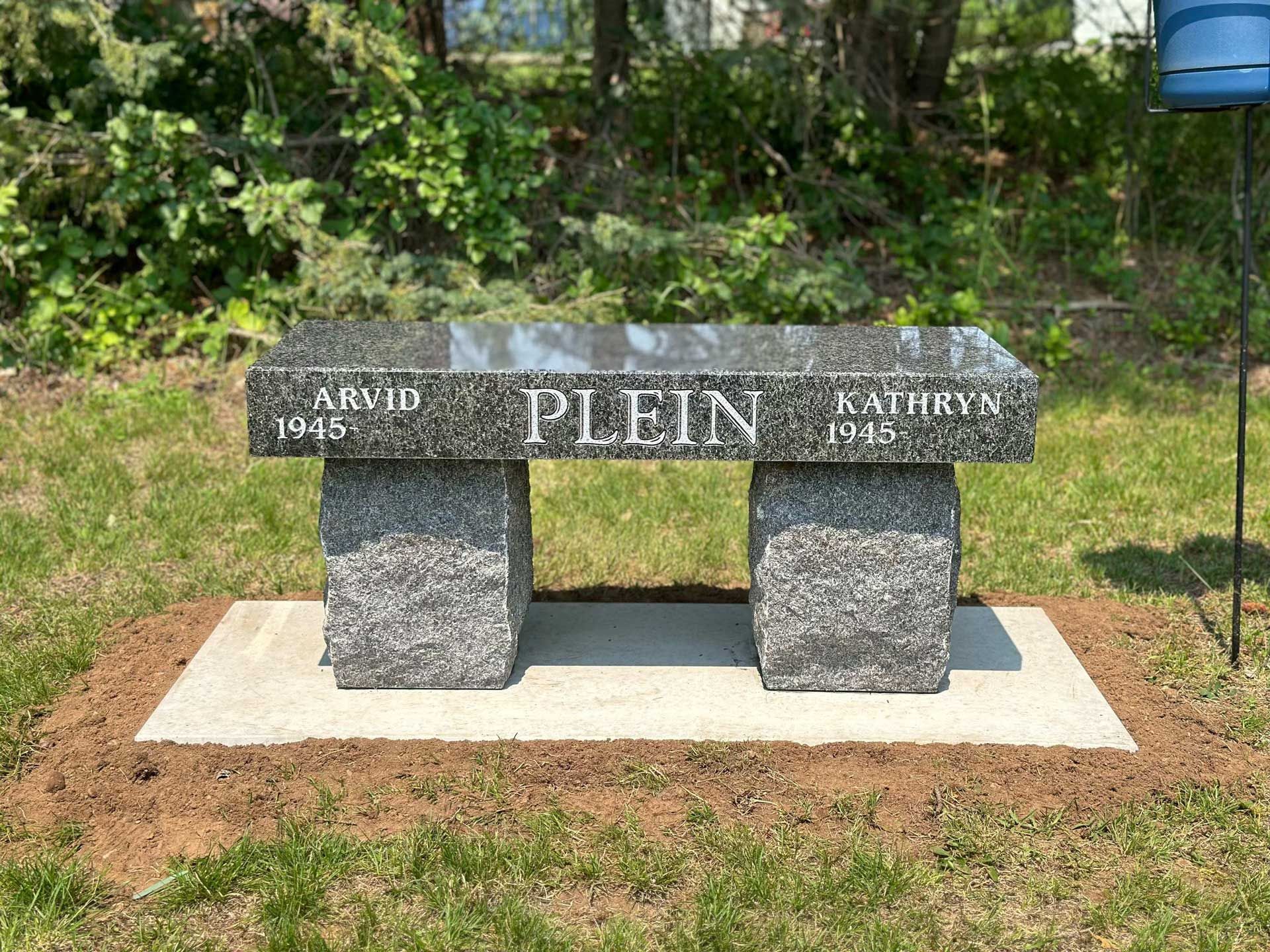A granite bench is sitting in the grass in a cemetery.