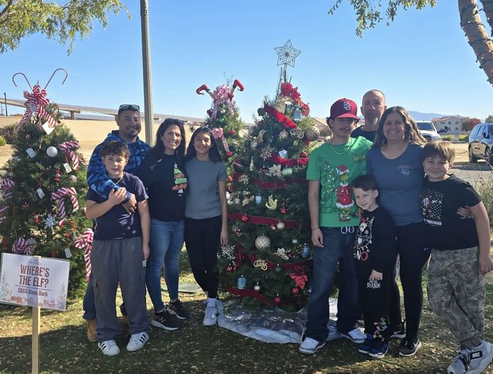 Group posing outdoors by decorated Christmas trees in a sunny park, with gifts and a festive holiday display.